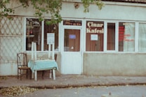 An exterior view of a small, slightly weathered caf&eacute; with signs on the windows and door, situated on a quiet street. The caf&eacute; is named '&Ccedil;ınar Kafesi'. A table with chairs and a light blue tablecloth sits outside, with upside-down chairs placed on top. A few leafy branches hang down from above.