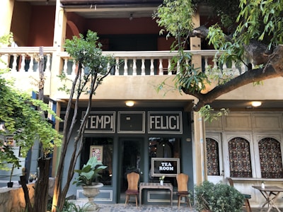A quaint tea room facade is surrounded by lush greenery and potted plants. The building features a balcony with ornate balusters and the words 'TEMPI FELICI' above the entrance. Two wooden chairs and a small table with a plant are placed outside, creating a cozy atmosphere.
