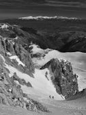 A mountainous landscape with snow-covered peaks and rocky cliffs, featuring two hikers walking through a snowy valley. The distant mountain range is visible under a partly cloudy sky, creating a sense of vastness and adventure.