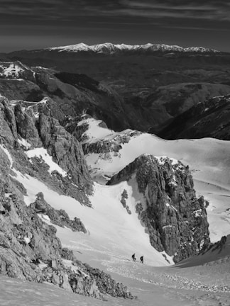 A mountainous landscape with snow-covered peaks and rocky cliffs, featuring two hikers walking through a snowy valley. The distant mountain range is visible under a partly cloudy sky, creating a sense of vastness and adventure.
