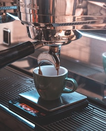 A close-up view of an espresso machine brewing coffee into a green ceramic cup placed on a metal drip tray. The deep brown coffee is flowing from the portafilter, and the machine's gleaming stainless steel body reflects light.