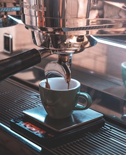 A close-up view of an espresso machine brewing coffee into a green ceramic cup placed on a metal drip tray. The deep brown coffee is flowing from the portafilter, and the machine's gleaming stainless steel body reflects light.