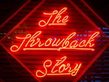 Bright red neon lights spell out 'The Throwback Story' on a background of wooden slats with a geometric pattern. The glow of the neon light creates a striking visual contrast against the dark background.