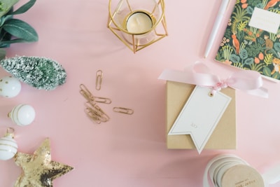 A neatly arranged flat lay featuring a variety of festive items on a soft pink surface. There is a small brown gift box wrapped with a pink ribbon and a white tag. Nearby is a gold geometric candle holder and a notebook with a botanical cover design. A group of gold paper clips and white baubles with gold accents are arranged to the left, alongside a small green decorative tree. A gold sequin star adds a sparkling touch.