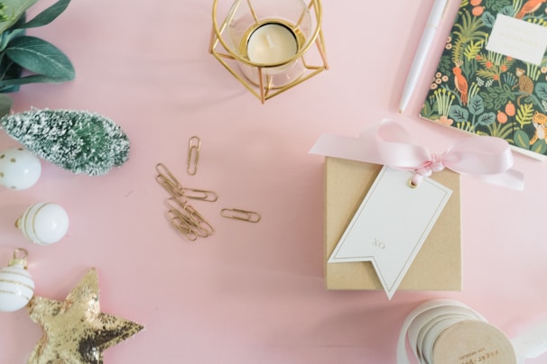 A neatly arranged flat lay featuring a variety of festive items on a soft pink surface. There is a small brown gift box wrapped with a pink ribbon and a white tag. Nearby is a gold geometric candle holder and a notebook with a botanical cover design. A group of gold paper clips and white baubles with gold accents are arranged to the left, alongside a small green decorative tree. A gold sequin star adds a sparkling touch.