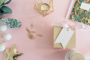 A neatly arranged flat lay featuring a variety of festive items on a soft pink surface. There is a small brown gift box wrapped with a pink ribbon and a white tag. Nearby is a gold geometric candle holder and a notebook with a botanical cover design. A group of gold paper clips and white baubles with gold accents are arranged to the left, alongside a small green decorative tree. A gold sequin star adds a sparkling touch.