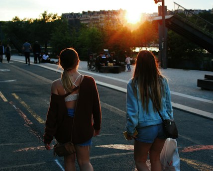Two people walk down a street toward a setting sun, surrounded by trees and distant buildings. The atmosphere is casual and summery, with one person wearing a brown sweater and shorts, and the other in a denim jacket and shorts. Other individuals can be seen interacting in the background.