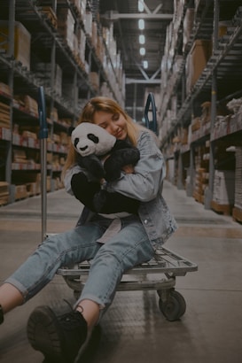 A young person wearing a denim jacket and jeans sits on a shopping cart in a warehouse aisle, hugging a plush panda toy. The warehouse shelves are stacked with boxes and products.