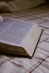 An open book lies on a plaid-patterned fabric surface, with visible text in a serif font. The soft lighting and focus on the pages create a calm atmosphere.