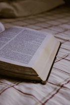An open book lies on a plaid-patterned fabric surface, with visible text in a serif font. The soft lighting and focus on the pages create a calm atmosphere.