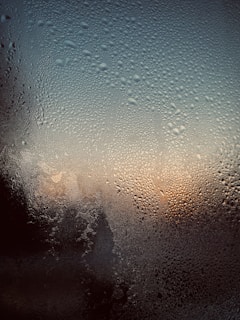 A close-up view of a window covered in condensation, with water droplets spread across the surface. The background suggests a sunset or sunrise, as warm orange and cool blue tones blend together.