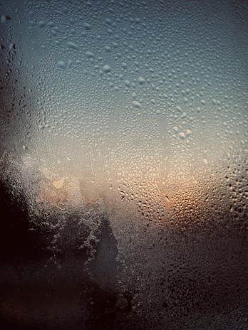 A close-up view of a window covered in condensation, with water droplets spread across the surface. The background suggests a sunset or sunrise, as warm orange and cool blue tones blend together.