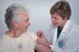 A healthcare professional in a white coat administers an injection to an older woman who is smiling. The woman is wearing glasses and earrings, and they are both focused on the procedure.
