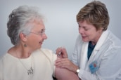 A healthcare professional in a white coat administers an injection to an older woman who is smiling. The woman is wearing glasses and earrings, and they are both focused on the procedure.
