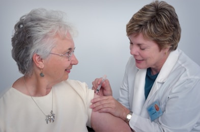 A healthcare professional in a white coat administers an injection to an older woman who is smiling. The woman is wearing glasses and earrings, and they are both focused on the procedure.