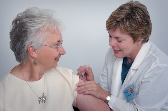A healthcare professional in a white coat administers an injection to an older woman who is smiling. The woman is wearing glasses and earrings, and they are both focused on the procedure.