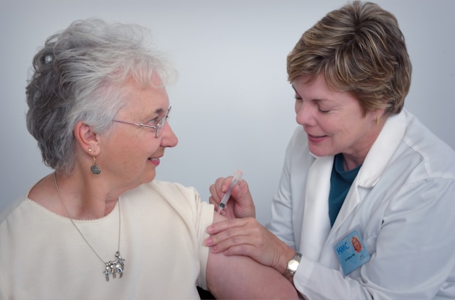 A healthcare professional in a white coat administers an injection to an older woman who is smiling. The woman is wearing glasses and earrings, and they are both focused on the procedure.
