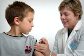 A young boy is receiving a vaccination from a smiling healthcare professional. The boy is looking away, while the healthcare worker gently administers the injection. Both are indoors with a plain background, suggesting a clinical setting.