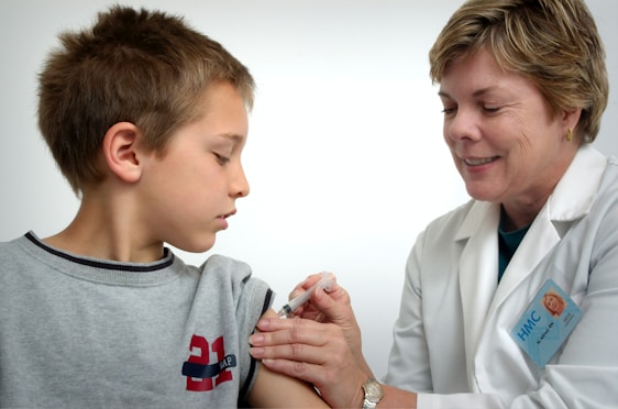 A young boy is receiving a vaccination from a smiling healthcare professional. The boy is looking away, while the healthcare worker gently administers the injection. Both are indoors with a plain background, suggesting a clinical setting.