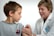 A young boy is receiving a vaccination from a smiling healthcare professional. The boy is looking away, while the healthcare worker gently administers the injection. Both are indoors with a plain background, suggesting a clinical setting.