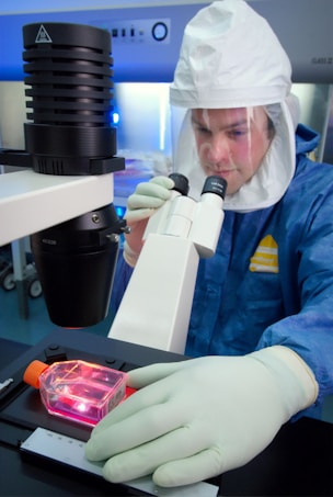 A person in protective clothing, including a hood and face shield, is using a microscope. Their hands are covered with pale green gloves, and they are observing a sample enclosed in a rectangular transparent container with a reddish content.