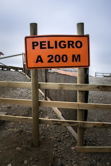 A wooden post supports an orange warning sign with black lettering that reads 'PELIGRO A 200 M'. The sign is situated in a rocky and gravelly area, with some fencing and terrain visible in the background under an overcast sky.