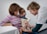 A healthcare professional is administering a vaccine to a young child while a woman, possibly the child's mother, holds the child on her lap. The child is focused on the procedure, and the healthcare provider is smiling, wearing a shirt with colorful heart patterns.