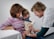 A healthcare professional is administering a vaccine to a young child while a woman, possibly the child's mother, holds the child on her lap. The child is focused on the procedure, and the healthcare provider is smiling, wearing a shirt with colorful heart patterns.