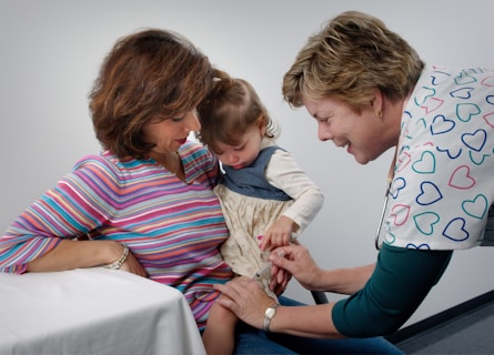 A healthcare professional is administering a vaccine to a young child while a woman, possibly the child's mother, holds the child on her lap. The child is focused on the procedure, and the healthcare provider is smiling, wearing a shirt with colorful heart patterns.