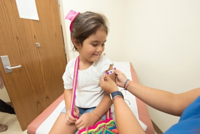 A young girl is seated on a medical examination table, receiving a small bandage on her arm from a healthcare provider. She is wearing a white shirt and has a pink bow in her hair. The provider is wearing bracelets and a blue shirt. The setting is a clinical room with wooden doors.