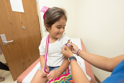 A young girl is seated on a medical examination table, receiving a small bandage on her arm from a healthcare provider. She is wearing a white shirt and has a pink bow in her hair. The provider is wearing bracelets and a blue shirt. The setting is a clinical room with wooden doors.
