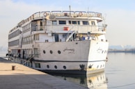 A large white cruise ship is docked at a pier. The ship has multiple decks with rows of windows and a variety of equipment on the top deck. It is secured to the dock with thick ropes. The background features a calm body of water and a distant shoreline with trees and buildings visible through a hazy sky.