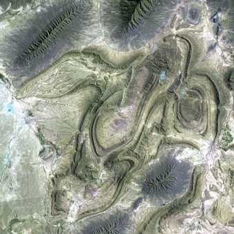 An aerial view of a dramatic and rugged landscape with ridges and valleys displaying intricate patterns. The image captures varying shades of green and brown, suggesting vegetation and bare earth. The topography features concentric circles and formations, possibly indicating geological activity or erosion over time.