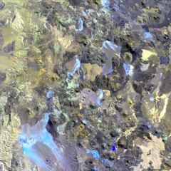Aerial view of a highly detailed geographical landscape, potentially a mountainous or volcanic region. Various water bodies in shades of blue are scattered across the terrain, surrounded by rugged hills, peaks, and valleys. The landscape features a mix of different textures, including rocky and potentially sandy areas, with a palette of colors ranging from earthy browns and greens to blues and grays.