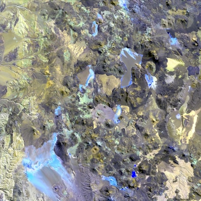 Aerial view of a highly detailed geographical landscape, potentially a mountainous or volcanic region. Various water bodies in shades of blue are scattered across the terrain, surrounded by rugged hills, peaks, and valleys. The landscape features a mix of different textures, including rocky and potentially sandy areas, with a palette of colors ranging from earthy browns and greens to blues and grays.