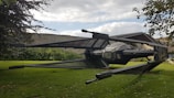 A large, detailed model of a spacecraft with angular wings and a central cockpit is positioned in a grassy garden area. Stone walls and lush greenery surround the setting, with tree branches partially visible in the foreground. The sky is partly cloudy, adding a dynamic backdrop.