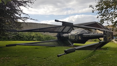A large, detailed model of a spacecraft with angular wings and a central cockpit is positioned in a grassy garden area. Stone walls and lush greenery surround the setting, with tree branches partially visible in the foreground. The sky is partly cloudy, adding a dynamic backdrop.
