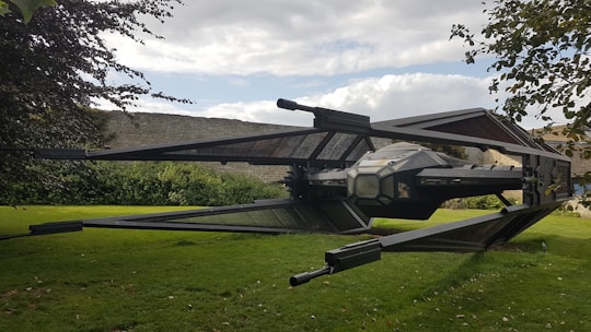 A large, detailed model of a spacecraft with angular wings and a central cockpit is positioned in a grassy garden area. Stone walls and lush greenery surround the setting, with tree branches partially visible in the foreground. The sky is partly cloudy, adding a dynamic backdrop.