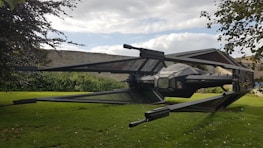 A large, detailed model of a spacecraft with angular wings and a central cockpit is positioned in a grassy garden area. Stone walls and lush greenery surround the setting, with tree branches partially visible in the foreground. The sky is partly cloudy, adding a dynamic backdrop.