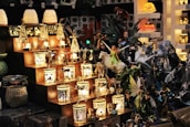 A collection of intricately designed lanterns displayed on wooden steps, surrounded by figurines of fairies and mythical creatures. The lights from the lanterns create a warm and inviting ambiance, highlighting the detailed craftsmanship of each item.