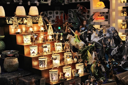A collection of intricately designed lanterns displayed on wooden steps, surrounded by figurines of fairies and mythical creatures. The lights from the lanterns create a warm and inviting ambiance, highlighting the detailed craftsmanship of each item.