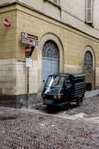 A small, three-wheeled vehicle is parked on a cobblestone street corner, adjacent to a beige-colored building with arched windows. Several signs indicate directions and display no-entry symbols.