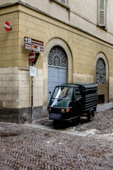 A small, three-wheeled vehicle is parked on a cobblestone street corner, adjacent to a beige-colored building with arched windows. Several signs indicate directions and display no-entry symbols.