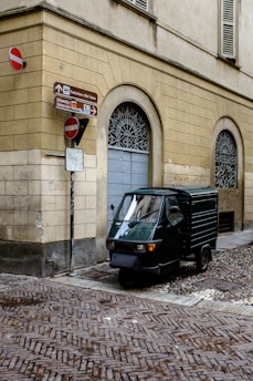 A small, three-wheeled vehicle is parked on a cobblestone street corner, adjacent to a beige-colored building with arched windows. Several signs indicate directions and display no-entry symbols.