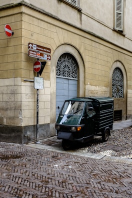 A small, three-wheeled vehicle is parked on a cobblestone street corner, adjacent to a beige-colored building with arched windows. Several signs indicate directions and display no-entry symbols.