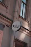 A circular sign with the letters K & B in black is mounted on an exterior wall of a building. The wall is light-colored with detailed architectural features such as molding and a drain pipe. The windows have dark frames and appear to be reflecting light.