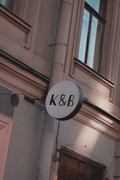 A circular sign with the letters K & B in black is mounted on an exterior wall of a building. The wall is light-colored with detailed architectural features such as molding and a drain pipe. The windows have dark frames and appear to be reflecting light.
