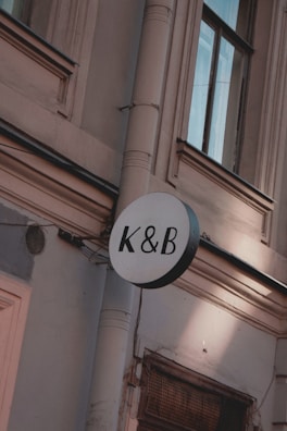 A circular sign with the letters K & B in black is mounted on an exterior wall of a building. The wall is light-colored with detailed architectural features such as molding and a drain pipe. The windows have dark frames and appear to be reflecting light.
