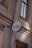 A circular sign with the letters K & B in black is mounted on an exterior wall of a building. The wall is light-colored with detailed architectural features such as molding and a drain pipe. The windows have dark frames and appear to be reflecting light.