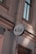 A circular sign with the letters K & B in black is mounted on an exterior wall of a building. The wall is light-colored with detailed architectural features such as molding and a drain pipe. The windows have dark frames and appear to be reflecting light.